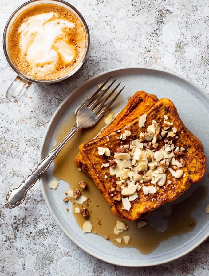 Three pieces of Pumpkin french toast in a blue plate with almond slices and maple syrup a latte is beside the dish