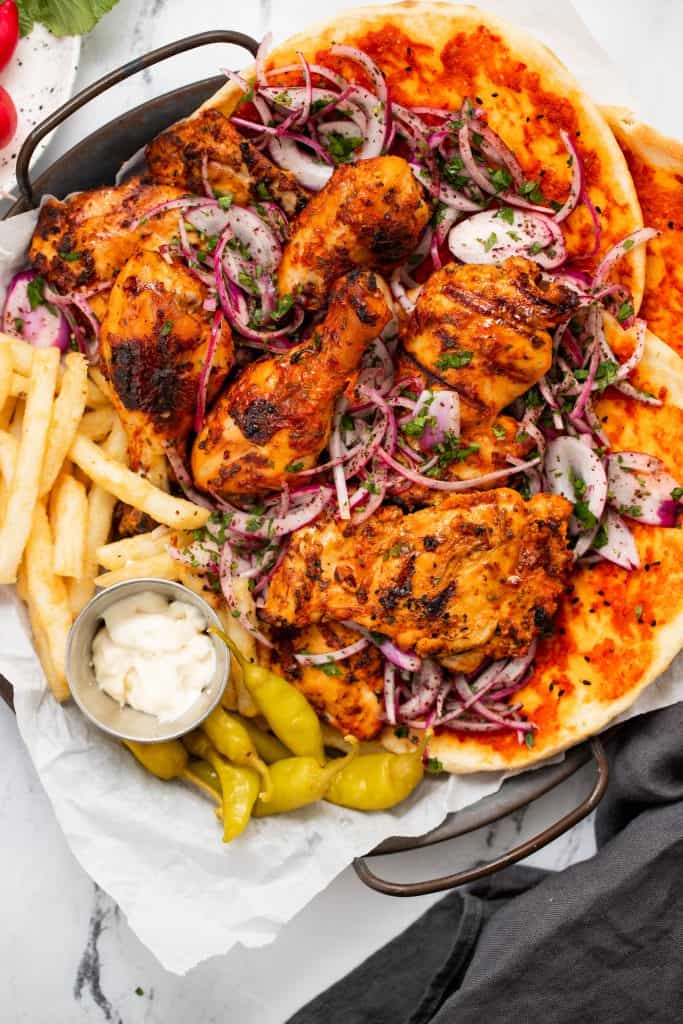 Close up of harissa BBQ chicken on a platter with fries, garlic mayo, sumac onions, and naan bread