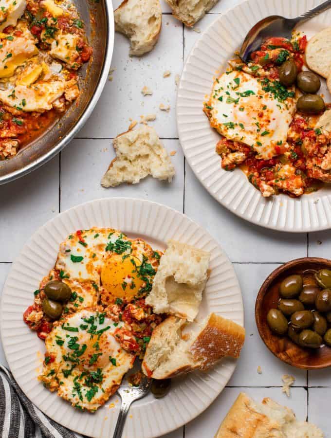 two plates fill with shakshuka, bead and olives side of olives bread and a pan with shakshuka