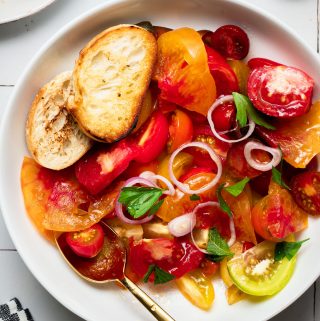 a white bowl filled with heirloom tomatoes, a golden serving spoon and a few pieces of bread